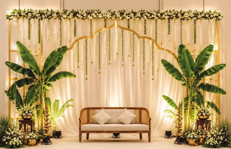 Cream draped wedding stage with a floral garland, cane sofa and brass lamps